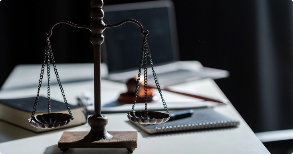 Bilancia della giustizia su una scrivania con martelletto, taccuino e laptop sullo sfondo.